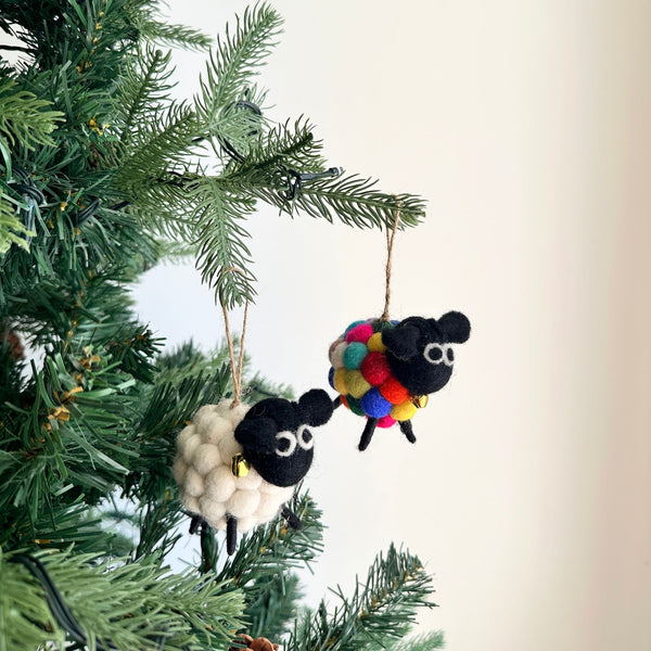 A set of two Mini Pompom Sheep felt Christmas ornaments hangs charmingly on a Christmas tree branch, presented against a bright, airy background. One white felt sheep has a black face, while the other black felt sheep is covered in colorful pompoms, both featuring a fluffy texture. Each whimsical sheep ornament includes a small golden bell around its neck and hangs from a jute string.