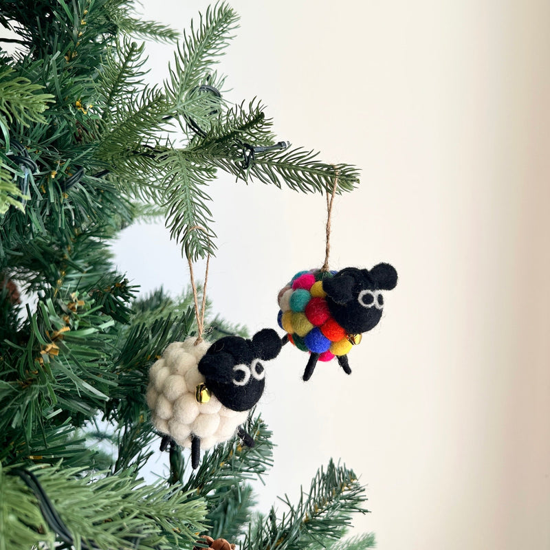 A set of two Mini Pompom Sheep felt Christmas ornaments hangs charmingly on a Christmas tree branch, presented against a bright, airy background. One white felt sheep has a black face, while the other black felt sheep is covered in colorful pompoms, both featuring a fluffy texture. Each whimsical sheep ornament includes a small golden bell around its neck and hangs from a jute string.