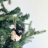 A fluffy white felt mini pompom sheep Christmas ornament, from a set of two, hangs on a vibrant green Christmas tree, radiating a festive and joyful mood. This charming handmade ornament features a black head with white eyes and small black ears, and a fluffy white body made of small felt balls. A tiny gold bell hangs around its neck, with a jute string attached for easy display.