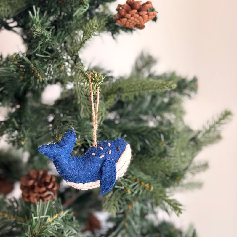 A deep blue felt whale Christmas ornament hangs from a branch of a softly blurred Christmas tree adorned with pine cones. This textured felt ornament features a light cream-colored underside and intricate white stitch accents, suspended by a jute string.