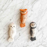 A set of three hand-stitched felt owl Christmas ornaments is displayed on a white marble surface. Each unique owl features a different color: one white with black stitches, one orange with white stitches, and one grey with white stitches. A red and white cotton string is attached to each for hanging.