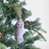 A whimsical hand-stitched felt owl ornament, part of a set of three felt Christmas ornaments, hangs on a festive green Christmas tree. This charming owl, crafted from light grey and lavender felt with visible stitching, features a round head, patterned body, and small black eyes. It is suspended by a red and white string, set against subtle tree lights and pine cones, creating a cozy holiday scene.