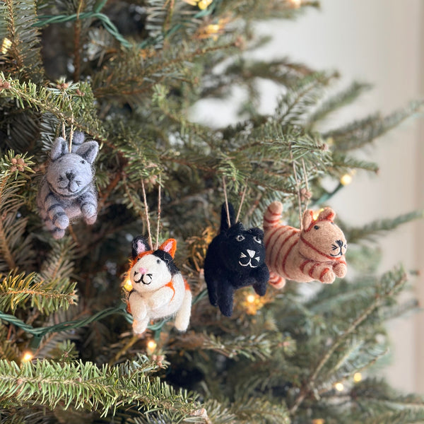A set of four felt cat Christmas ornaments hangs among the green needles of a Christmas tree, illuminated by small, warm lights. Crafted from 100% felt, the unique Christmas ornaments include a gray striped cat, a white cat with black and orange spots, a black cat, and a tan striped cat. Each features pointy ears, a tail, and stitched markings, suspended by a jute string.