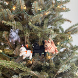 A set of four felt cat Christmas ornaments hang on a festive Christmas tree, glowing with warm decorative lights. Each handcrafted felt cat ornament, suspended by a jute string, features unique colors and patterns, including gray striped, white with black and orange, solid black, and light brown striped. Small details like ears, tails, and embroidered facial features add a whimsical touch to these festive Christmas tree ornaments.