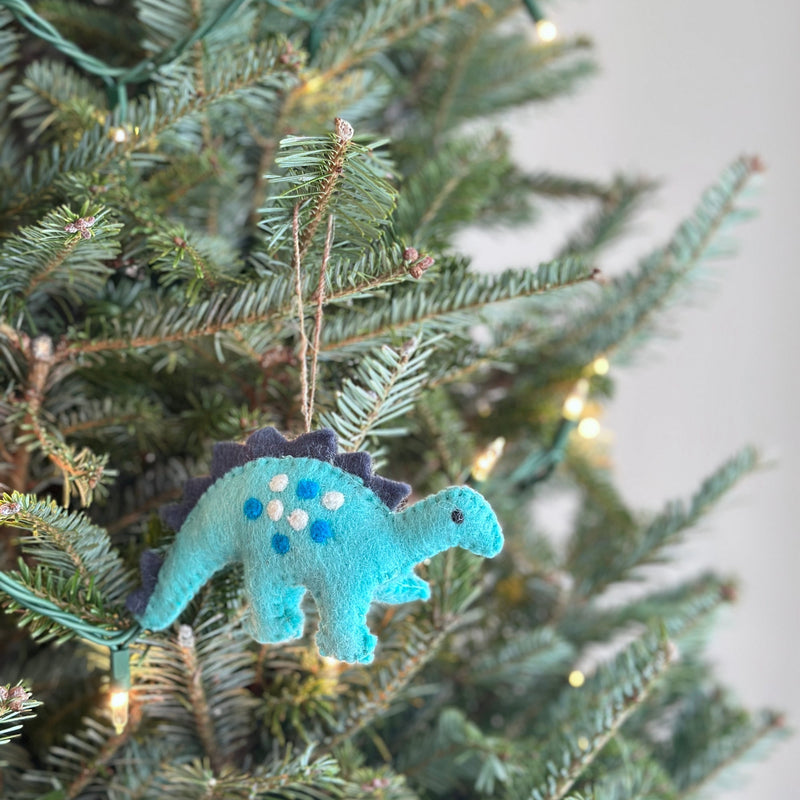 A charming felt Christmas dinosaur ornament is displayed on a vibrant Christmas tree adorned with warm, glowing lights. This light blue felt dinosaur features darker blue accents along its spine and playful white and blue polka dots on its side. A jute string is attached for easy hanging, blending with the cozy holiday atmosphere.