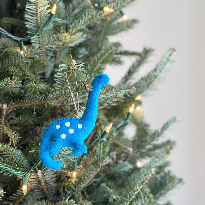 A bright blue felt dinosaur ornament, part of a Christmas set, hangs gracefully from a lush Christmas tree branch. This detailed felt ornament features a long neck, small head, four short legs, and white spots. A jute string is attached for hanging, with warm white lights on the tree enhancing the festive mood.