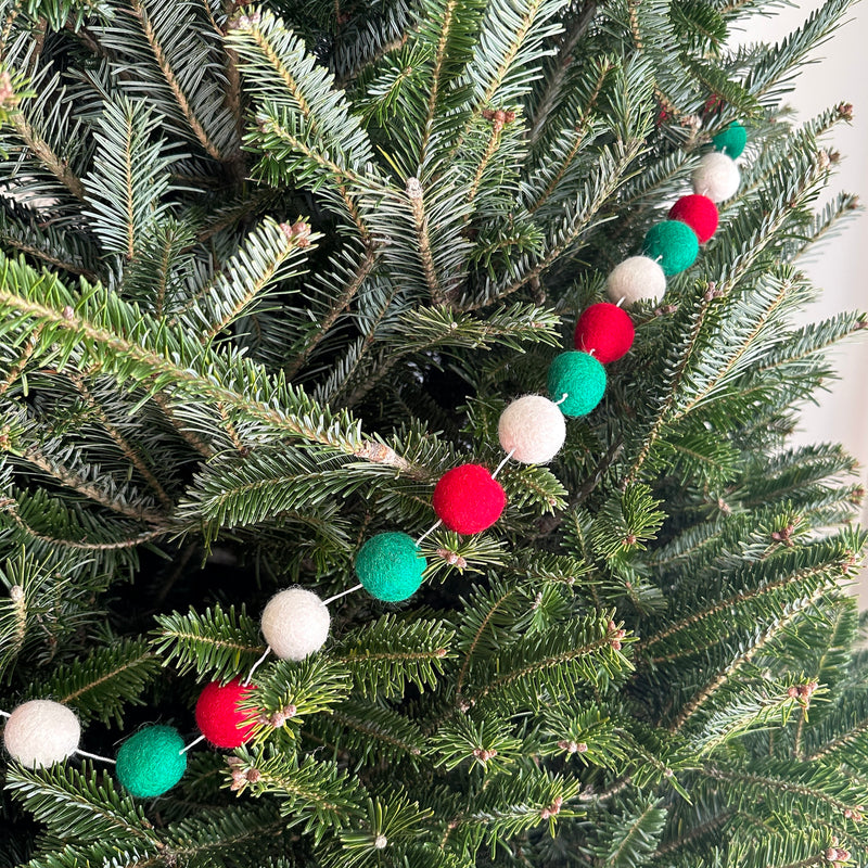A festive Felt Christmas Pompom Ball Garland drapes over the green branches of a Christmas tree. The felt garland features a classic pattern of alternating red, white, and green pompom balls strung on a thin white string, creating a cozy holiday ambiance.