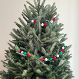 A felt Christmas pompom ball garland hangs gracefully on a decorated Christmas tree. The vibrant garland features spherical felt pompom balls in a repeating pattern of red, green, and white. This festive decoration adds a playful yet elegant touch, creating a warm, inviting holiday atmosphere.