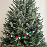 A festive felt Christmas Pompom Ball Garland is draped horizontally across a section of a lush green Christmas tree. The garland features smooth, round felt balls in alternating red, white, and green, evenly spaced along a string. It brings a cheerful holiday mood and classic Christmas charm, enhancing the elegance of holiday decorating.