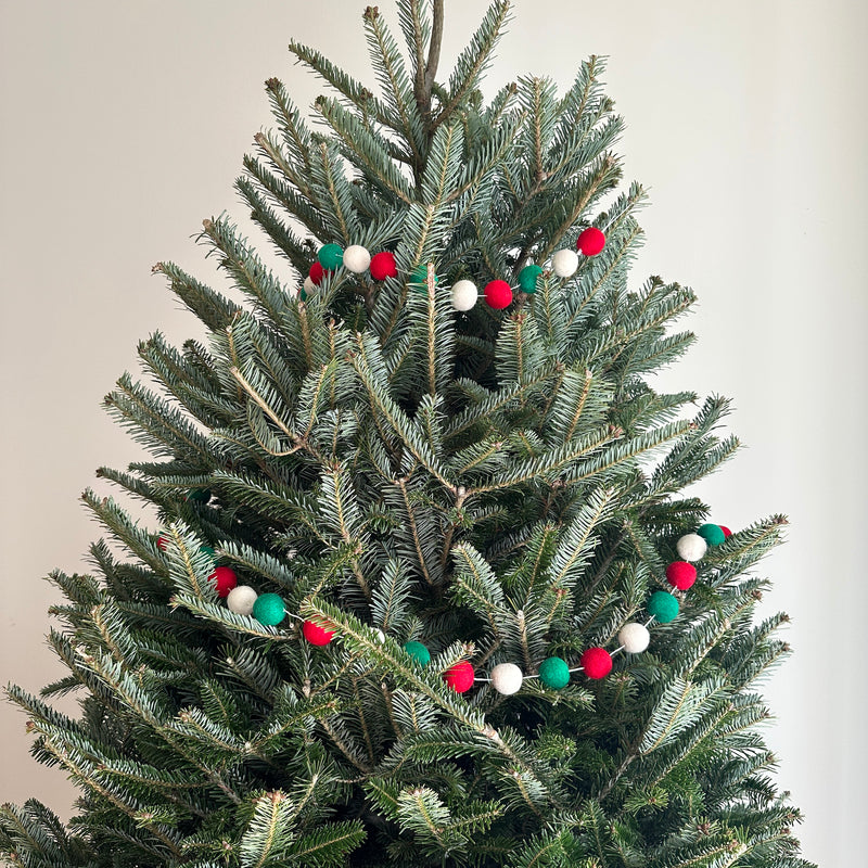 A festive Felt Christmas Pompom Ball Garland is draped around the branches of a Christmas tree, creating a playful holiday atmosphere. The garland features spherical felt pompom balls in red, green, and white, evenly spaced on a string.