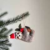 A gray felt sloth Christmas ornament hangs from a tree branch against a plain white background. The charming handmade felt sloth wears a red and white Santa hat and holds a festive branch with green leaves and red berries.