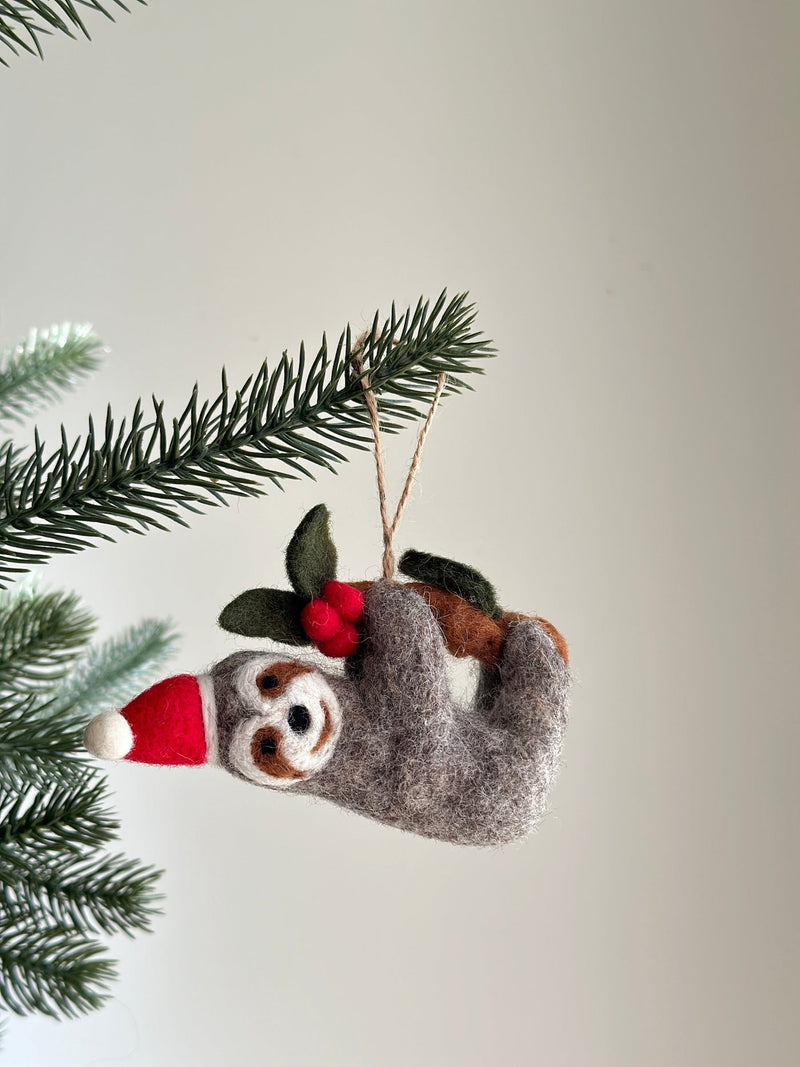 A felt Christmas sloth ornament hangs from a Christmas tree branch. This charming felted sloth wears a red Santa hat with a white pompom and holds a small brown felt branch adorned with green leaves and red berries. The handcrafted ornament hangs by a jute string.