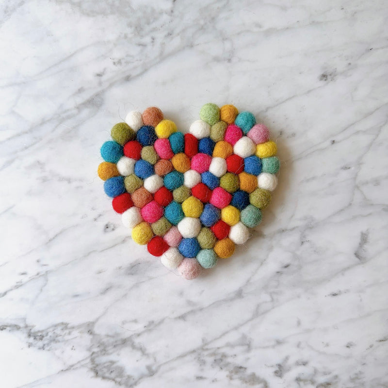 A single heart-shaped rainbow felt cup coaster, part of a set, rests on a bright white marble surface. This unique handmade felt coaster is crafted from tightly packed, hand-stitched felt balls made of 100% organic lamb wool. The colorful design features white, red, yellow, blue, and green felt balls, forming a cheerful rainbow pattern.