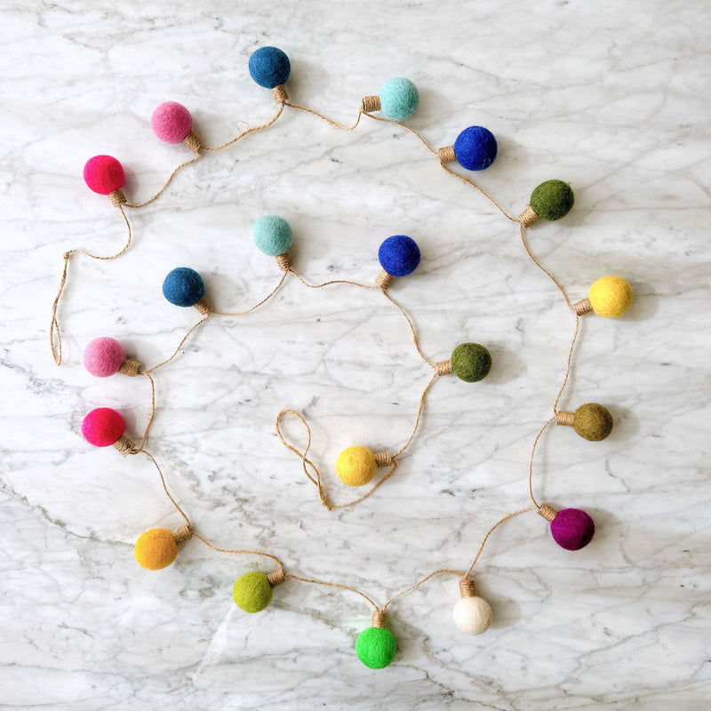 A colorful felt lightbulb garland is displayed in a curved pattern on a clean, elegant marbled surface. This festive wool felt garland features spherical felt balls, each resembling a lightbulb with a twine-wrapped stem, attached to a light brown twine. The vibrant felt 'lightbulbs' come in red, pink, blue, light blue, green, yellow, purple, and white, adding a cheerful touch.