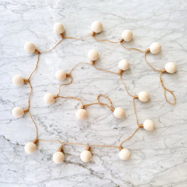 A wool felt lightbulb garland rests on a light gray marble surface, viewed from above. This felt color ball lightbulb garland features spherical, off-white wool felt balls that mimic lightbulbs. Each 'lightbulb' is secured with a thin, rustic brown string, creating a minimalist and appealing design with neutral tones.