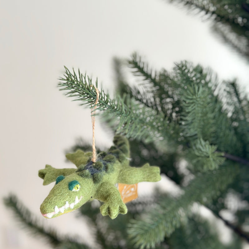A felted wool gator ornament, designed to appear crawling, hangs from a vibrant green Christmas tree. This unique Christmas tree ornament features a textured body with meticulously crafted details like its eyes, teeth, and back. A jute string allows the handcrafted felt gator to hang gracefully.