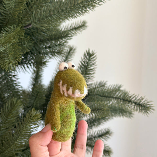 A handmade wool felt gator finger puppet is held in a female hand, set against a festive green Christmas pine tree and a plain white wall. The cheerful gator puppet is predominantly green with a lighter green belly, small white eyes, and visible white teeth. This wool felt piece showcases a textured surface and small, stubby arms, highlighting its handcrafted detail.