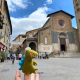 A cute, ethically handcrafted wool felt gator finger puppet is playfully held by a hand in a lively outdoor town square, featuring vintage buildings and a cathedral. This compact, predominantly green gator puppet showcases blue fins, small white eyes, and a tiny pink tongue, reflecting its unique and whimsical design for imaginative play.