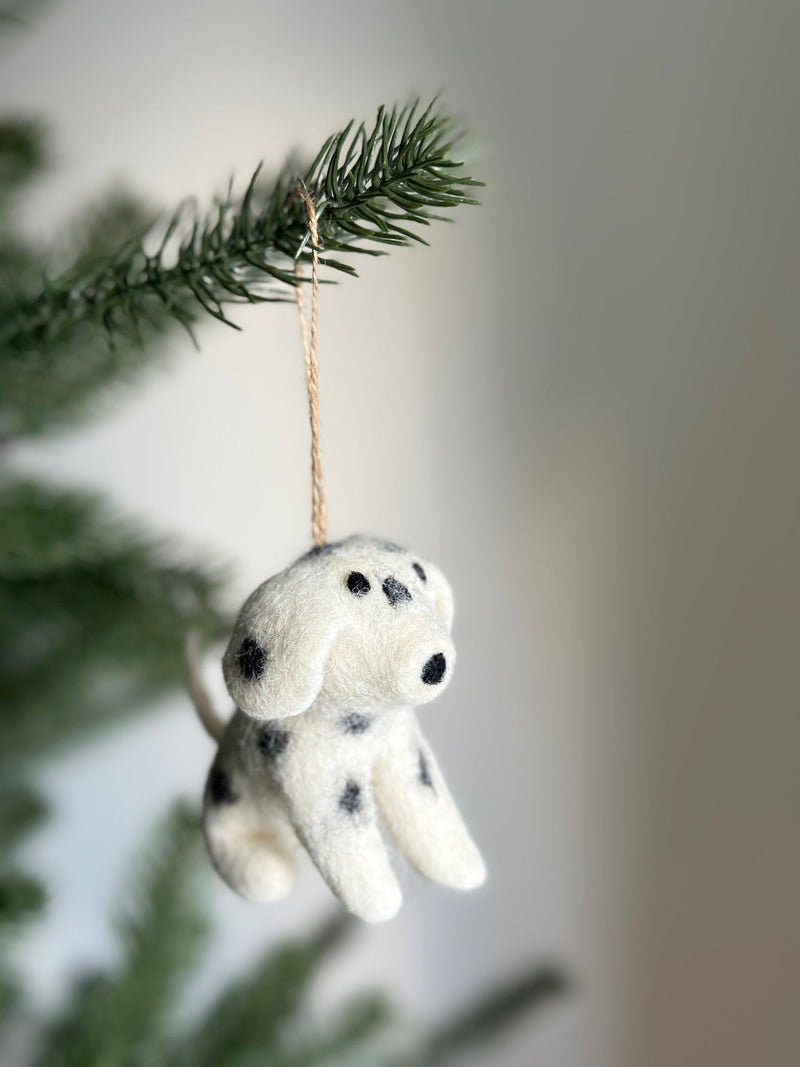 A charming Felt Dalmatian Ornament hangs from a Christmas tree branch, suspended by a jute string. This unique Christmas ornament portrays a playful Dalmatian pup crafted from felt, depicted in a sitting position with distinctive black spots, a small tail, and ears.