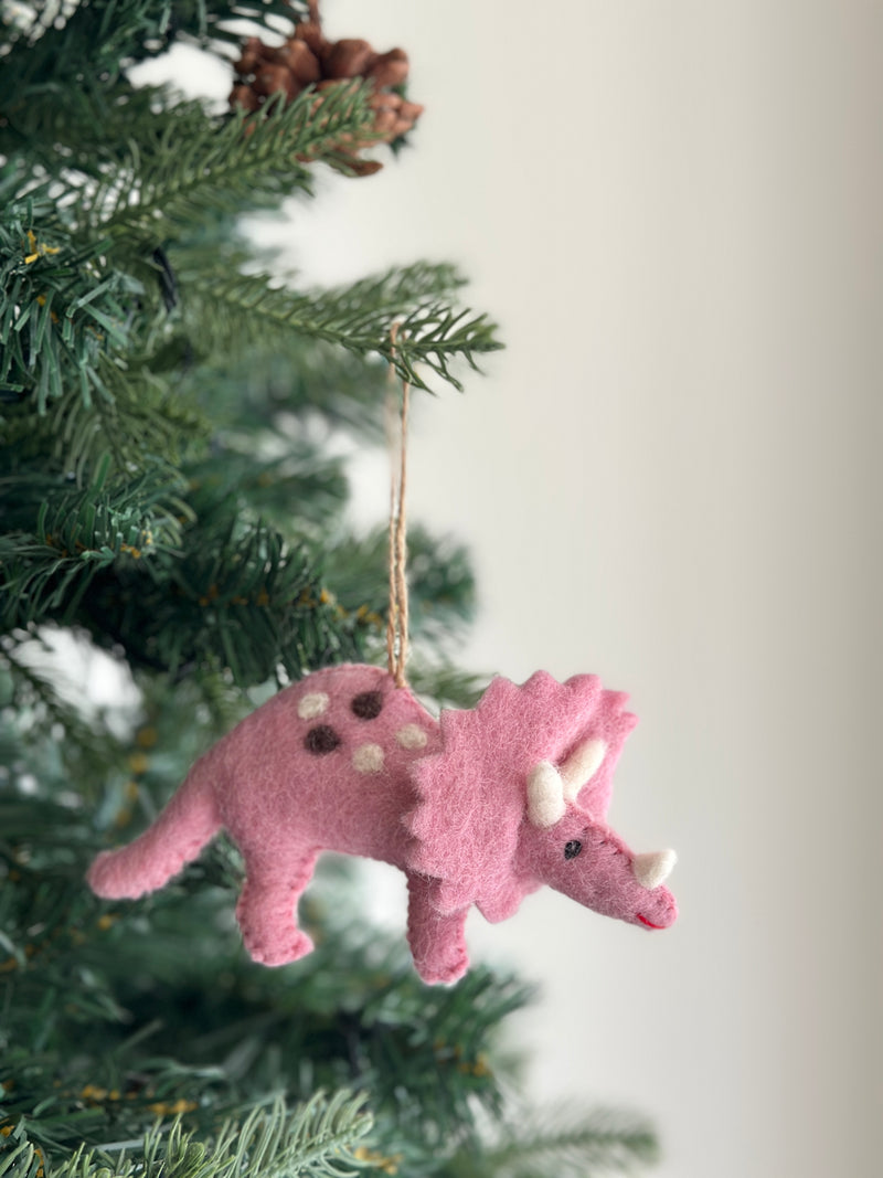 A pink triceratops felt dinosaur ornament hangs on a vibrant green Christmas tree. This unique Christmas ornament, crafted from wool felt, features brown and white spots, a white horn, and a red smiling mouth. A jute string is visible for hanging, with lush green pine needles and a pine cone in the background, creating a festive scene.