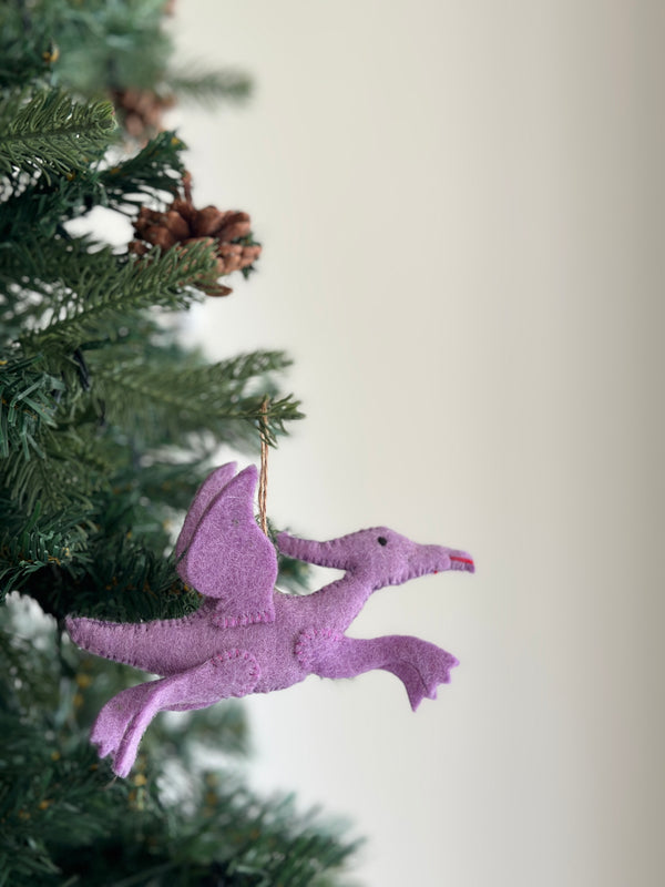 A vibrant purple pterosaur felt dinosaur ornament is suspended by a jute string on a Christmas tree, nestled among green needles and pine cones. This wool felt ornament appears to be in mid-flight, showcasing hand-stitched edges, small black eyes, and a red beak. Its whimsical design adds a charming touch to the festive decor.
