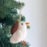 A handcrafted wool felt dove Christmas tree ornament hangs from a lush green branch, radiating festive charm against a soft, off-white backdrop. This pure white dove ornament features a fluffy body, an orange beak and feet, and delicately holds an olive twig in its beak. It includes a jute string for easy hanging.