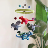 A felt dragon baby mobile hangs in front of a cream-colored wall and a leafy green plant, creating a whimsical scene. It features a light green felt ring from which colorful felt balls and several felt dragons are suspended. The dragons, in shades of red, dark gray, green, and blue, hang at different heights, each detailed with contrasting wings and horns.