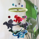 A whimsical felt dragon baby mobile is suspended indoors against a lush green plant backdrop. It features red, blue, green, and gray felt dragons hanging from a round, light-green felt ring by thin white strings. Colorful felt balls are attached above the ring, adding to its playful charm.