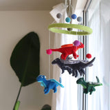 A felt dragon baby mobile hangs in front of a window, showcasing a whimsical design. It features a circular green felt frame adorned with gray, blue, and green felt balls. Four colorful felt dragons are suspended on white strings: a red one with orange wings, a dark gray one with lighter accents, a blue one with lighter wings, and a green one with white wings. A large green leaf partially obscures the left side of the mobile.