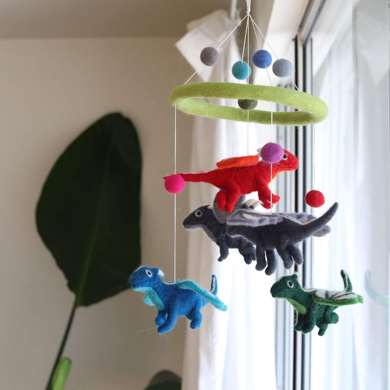 A felt dragon baby mobile hangs in front of a window, showcasing a whimsical design. It features a circular green felt frame adorned with gray, blue, and green felt balls. Four colorful felt dragons are suspended on white strings: a red one with orange wings, a dark gray one with lighter accents, a blue one with lighter wings, and a green one with white wings. A large green leaf partially obscures the left side of the mobile.