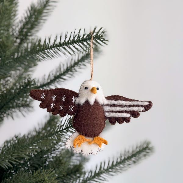 A patriotic felt eagle ornament hangs from a green pine branch, resembling a Christmas tree, against a clean white backdrop. This unique Christmas tree ornament features a brown felt body, white felt head and tail, a yellow felt beak with dark eyes, and orange felt feet. Its wings are detailed with stitched white stars and alternating brown and white stripes, showcasing an American spirit. A jute string allows for easy hanging.