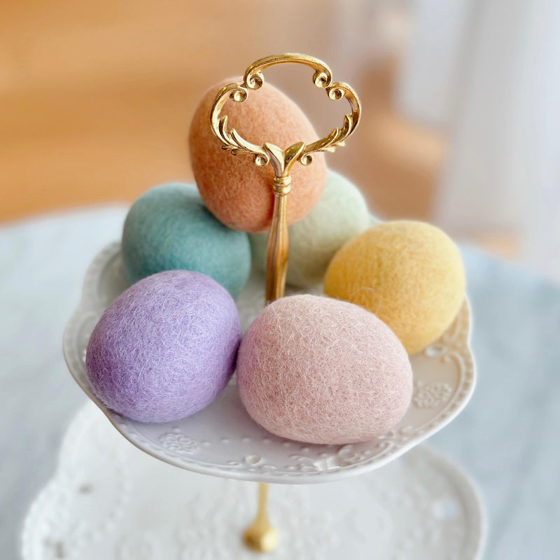 Wool felt Easter eggs rest on a two-tiered white serving stand with a golden handle and lace pattern, set against a soft, blurred background. The set includes six life-sized, wool felt Easter eggs in delicate pastel, macaron-inspired colors: peach, light green, yellow, blue, pink, and lavender. Each felt Easter egg has a textured surface, evoking springtime charm.