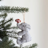 A gray wool felt elephant Christmas ornament, playful and handcrafted, hangs from a festive Christmas tree branch. The charming elephant is seated, with front legs slightly raised, and its trunk holds a brown football with white stripes. A small black eye and a jute string for hanging complete this whimsical, unique Christmas ornament.
