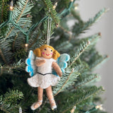 A Felt Fairy Girl Ornament hangs on a lush green Christmas tree, adorned with small, warm white lights, creating a festive and joyful atmosphere. The handcrafted felt ornament features a tan skin tone, rosy cheeks, and curly golden hair. She wears a short, white dress with delicate floral accents and a black belt. Her wings have three white fluffy scallops with light blue accents, and she sports white ballet shoes with crisscrossed laces, ready for dancing arrangements.