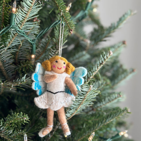 A Felt Fairy Girl Ornament hangs on a lush green Christmas tree, adorned with small, warm white lights, creating a festive and joyful atmosphere. The handcrafted felt ornament features a tan skin tone, rosy cheeks, and curly golden hair. She wears a short, white dress with delicate floral accents and a black belt. Her wings have three white fluffy scallops with light blue accents, and she sports white ballet shoes with crisscrossed laces, ready for dancing arrangements.