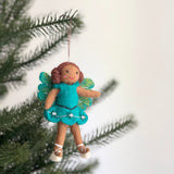 A felt fairy girl ornament hangs gracefully from a lush green Christmas tree, set against a soft white backdrop. The handmade felt ornament features a fairy with brown skin, curly brown hair, and a cheerful red smile. She is dressed in a bright turquoise felt dress with clear beads at the waist and has translucent green and blue wings. A red cotton string suspends this enchanting Christmas tree ornament.