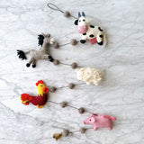 A playful wool felt farmland animal garland is displayed on a marbled white surface. The decorative garland features a gray felt horse, a white and black felt cow, a white felt sheep, a red and yellow felt rooster, and a pink felt pig. Small brown wool balls connect the felt animals, with a copper bell at one end, creating a whimsical and rustic piece.