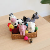 A wool felt farm animal garland rests in a woven basket on a light wooden table, bathed in soft, natural light. This charming nursery garland features needle-felted 3D figures: a white and black cow with a pink snout, a gray horse, a pink pig, a white sheep, and a red and yellow rooster. A small copper bell hangs at the end.