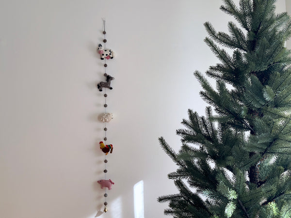 A felt farmland animal garland hangs on a white wall beside a green Christmas tree, creating a festive atmosphere. This 4-foot handmade wool felt garland features five detailed 3D needle-felted animals: a black and white milk cow, a grey horse, a white sheep, a red rooster, and a pink pig. Grey felt balls separate the animals along the string, which ends with a copper bell.