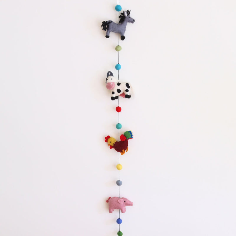 A playful felt farm animal garland hangs vertically against a plain white background. It features a grey wool felt horse with a black mane, a white cow with black spots and a pink snout, a rooster with a red head and tail feathers, and a pink pig. Colorful felt balls are strung between the needle-felted animals, with a single copper bell at the bottom.