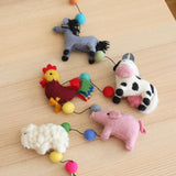 A Felt Farm Animal Garland, approximately 4-foot long, is playfully displayed on a light wooden table. This handcrafted, three-dimensional 100% wool felt garland features a gray horse with black mane, a rooster with red and yellow feathers, a white and black spotted milk cow, a pink pig, and a fluffy white sheep. Colorful felt balls in shades of orange, green, blue, yellow, pink, and gray are interspersed, and a copper bell hangs at one end.