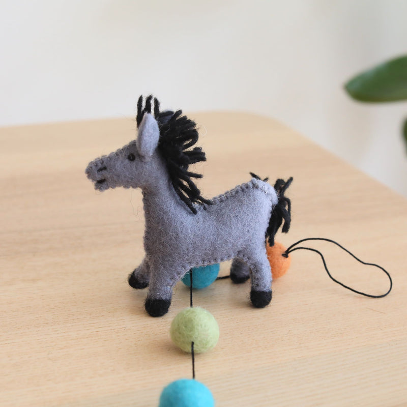 A single light gray felt horse, part of a wool felt farm animal garland, rests on a wooden table against a soft white background. The horse features a light gray body with black hooves, mane, and tail. Its needle-felted wool texture gives it a soft, handcrafted appearance. It is connected to a string of round wool felted balls in teal, light green, and orange, and includes a small bell at one end.