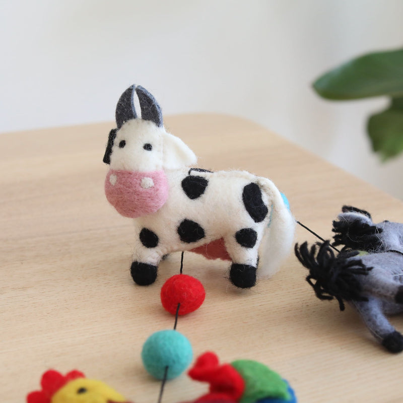 A wool felt farmland animal garland is displayed on a light brown wooden surface. This charming handmade garland features 3D needle-felted animals, including a white milk cow with black spots, a pink snout, and gray ears, a gray horse, and a rooster. The figures are suspended by black strings, ending with a copper bell. A blurred green plant is in the background.