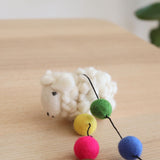 A wool felt farm animal garland features a fluffy, off-white felt sheep resting on a wooden table against a neutral background. The sheep's soft texture and small stitched eyes and nose highlight its handcrafted charm, creating a calm, natural display.