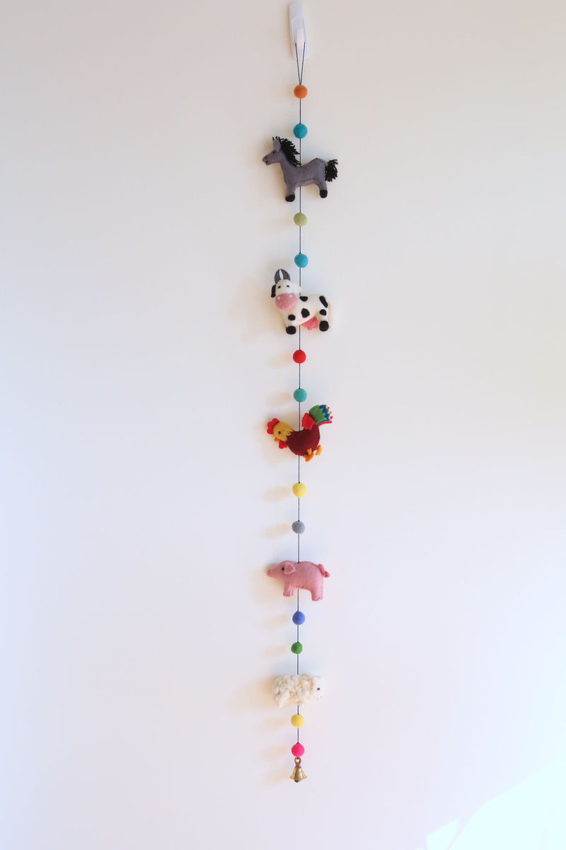 A felt farmland animal garland hangs playfully against a plain white wall, featuring five 3D felted figures. The decorative garland includes a gray horse, a black and white milk cow, a red and yellow rooster, a pink pig, and a white sheep, with a copper bell at its end.