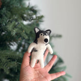 A handcrafted wool felt Alaskan husky dog finger puppet, held by a female hand, is set against a softly blurred Christmas tree background. This unique finger puppet showcases intricate needle felt detailing, including black markings around its eyes, a small black nose, and prominent erect triangular ears.