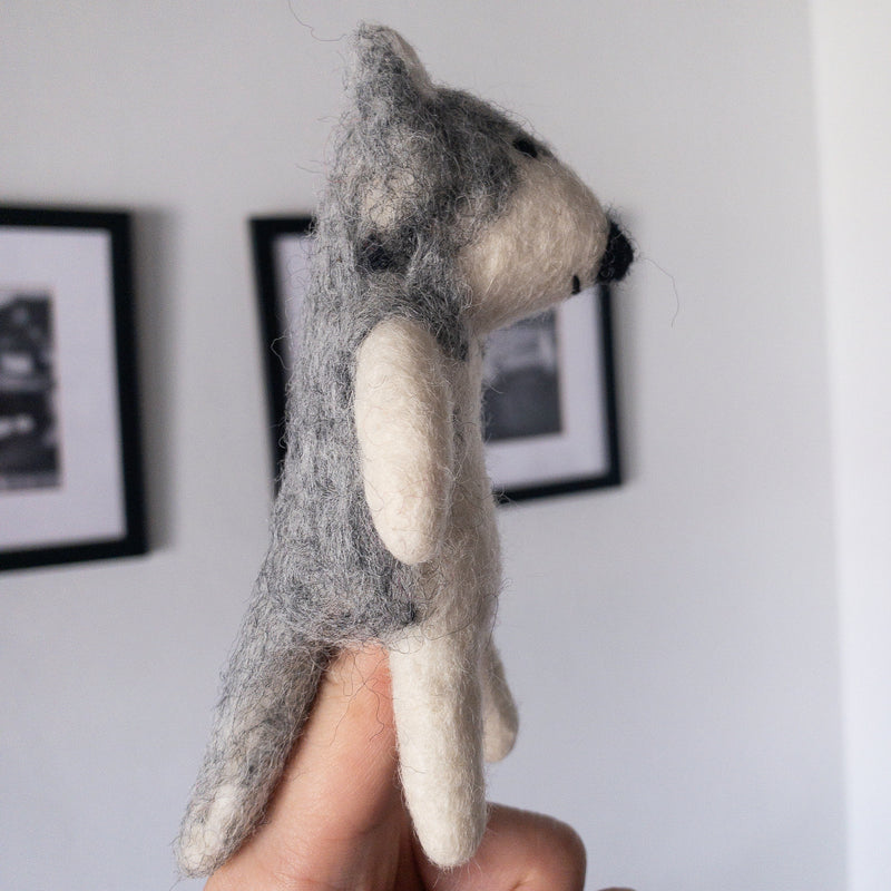 An Alaskan Husky dog felt finger puppet is held by a female hand against a softly lit, minimalist white background with framed pictures. This handmade wool felt finger puppet, crafted with intricate needle felt detailing, features a mix of gray and white. It captures the husky's fluffy texture, erect triangular ears, and defined shape, showcasing its playful, handcrafted charm.