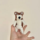 A handcrafted wool felt Alaskan husky dog finger puppet rests gently on a model's open hand. This needle-felted puppet is primarily white with brown markings on its head and body, featuring small triangular ears and a black nose. Simple stitched details give it a playful, smiling expression, capturing the Alaskan Husky's distinctive look.