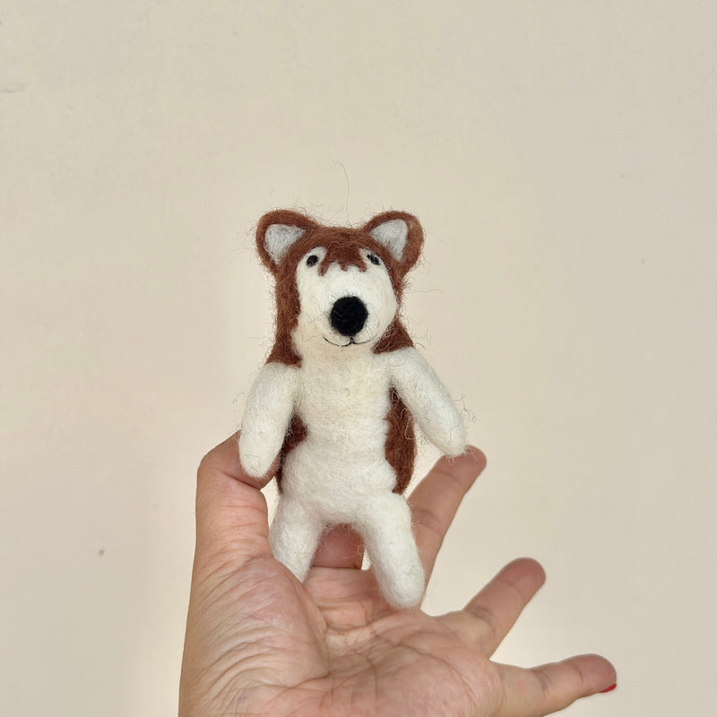 A handcrafted wool felt Alaskan husky dog finger puppet rests gently on a model's open hand. This needle-felted puppet is primarily white with brown markings on its head and body, featuring small triangular ears and a black nose. Simple stitched details give it a playful, smiling expression, capturing the Alaskan Husky's distinctive look.