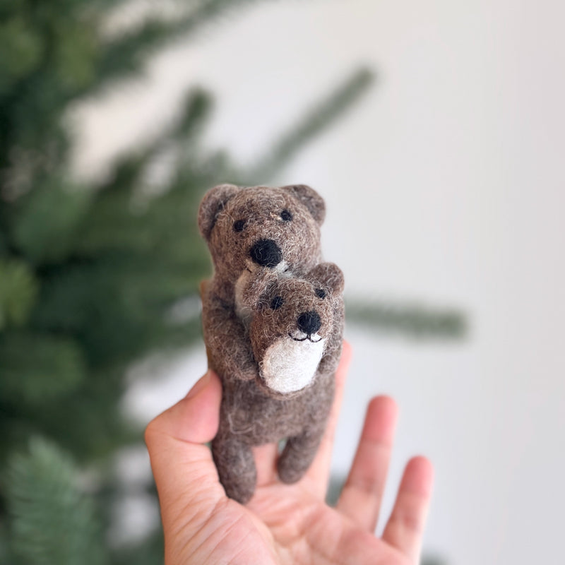 A handmade felt American River Otter finger puppet, featuring a mother otter cradling her baby, is held in a female hand. The brown felt mother otter gently holds her smaller baby, which has a white belly. This textured felt toy is presented against a blurred Christmas tree background, highlighting its handcrafted warmth for imaginative play.